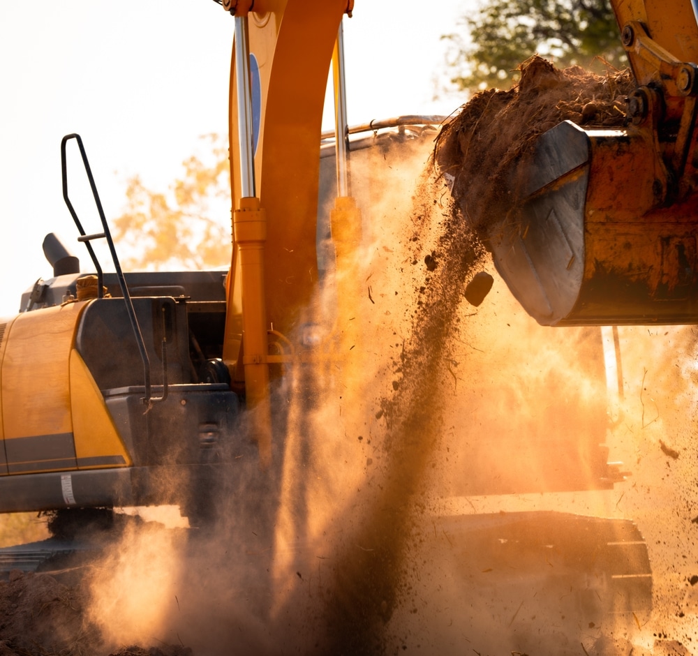 Excavatrice jaune déversant de la terre dans un nuage de poussière sous une lumière dorée de fin de journée.