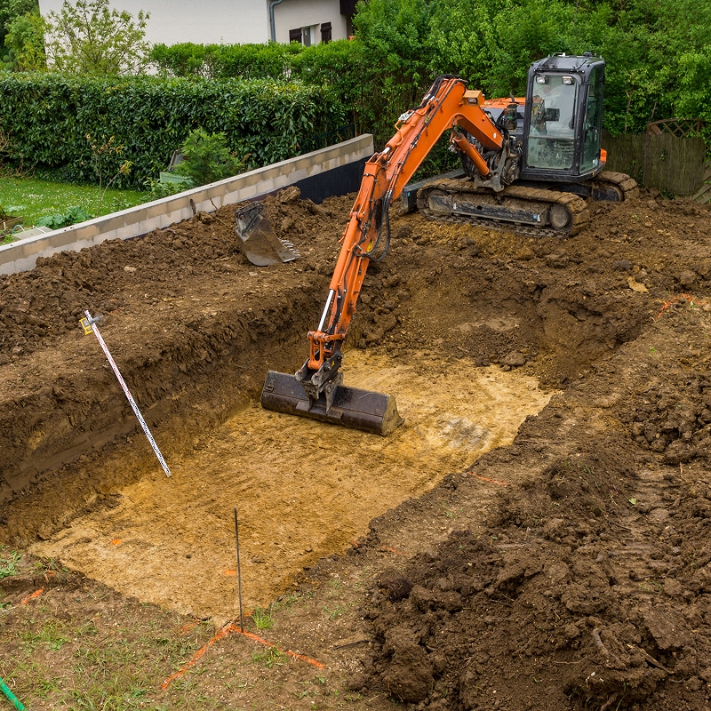 Une pelleteuse orange creuse une fosse rectangulaire pour des fondations dans un jardin avec une mire de nivellement.