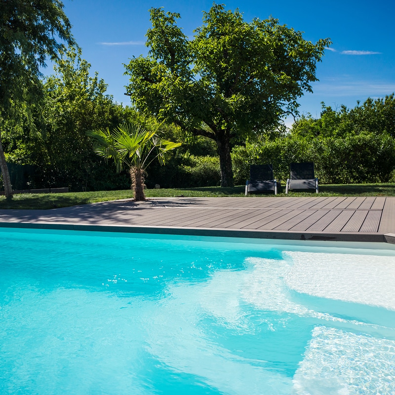 Piscine enterrée aux eaux turquoise avec terrasse en bois, palmier et chaises longues sous un ciel bleu dégagé.