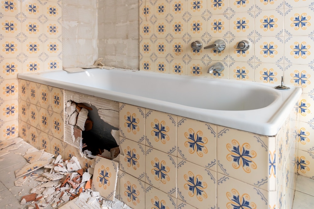 Rénovation de baignoire : réparation de carrelage cassé Baignoire en rénovation avec carreaux cassés aux motifs floraux bleus et jaunes et débris au sol.